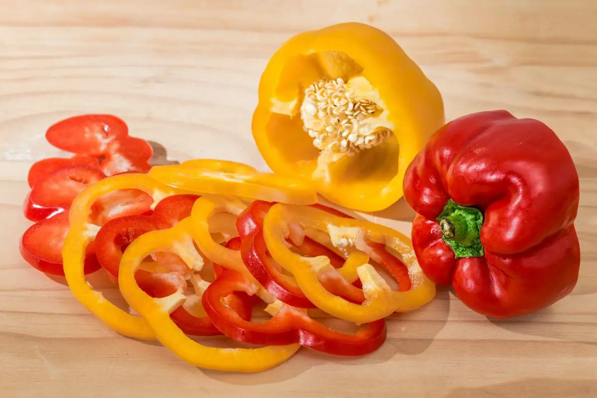 A sliced yellow and red bell pepper lie on a light wooden surface, with some slices in front and whole and partially sliced peppers behind.