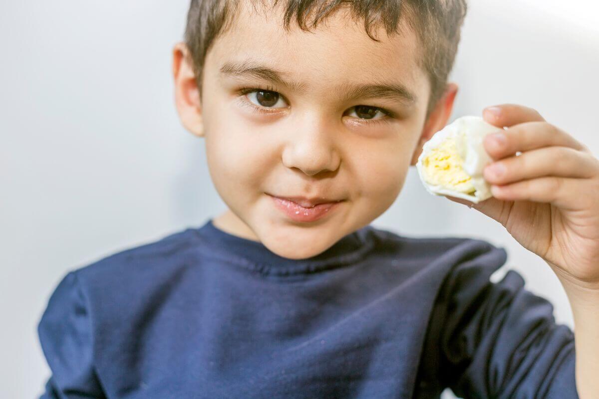 A young child with short dark hair is holding up a half-eaten hard-boiled egg towards the camera. 