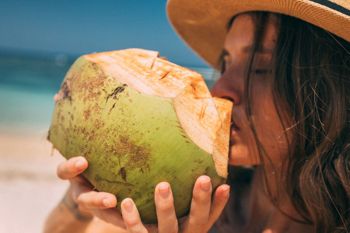 A person wearing a straw hat drinks from a freshly cut green coconut on a sunny beach. 