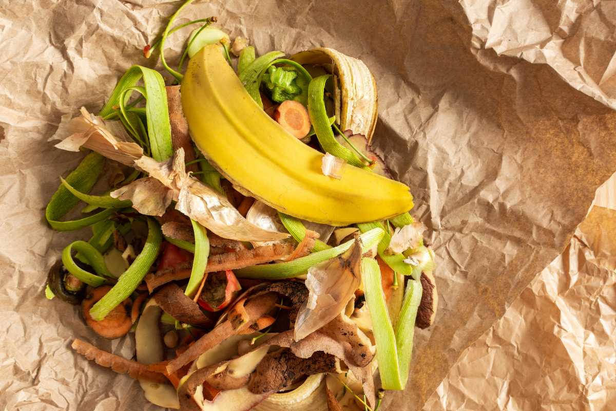 A pile of assorted vegetable and fruit peels, including a banana peel, carrot peels, and other organic scraps, sits on crumpled brown paper.