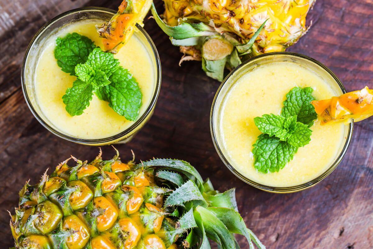 Two glasses of pineapple smoothie garnished with mint leaves and pineapple slices are placed on a wooden surface.