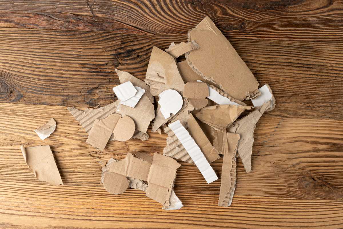 A pile of ripped and torn pieces of corrugated cardboard is scattered on a textured wooden surface. The various shapes and sizes reveal visible ridges with some white inner layers exposed.