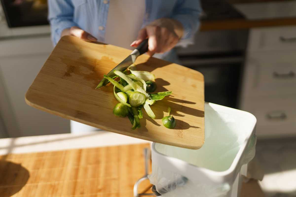 A person empties vegetable peels and scraps from a wooden cutting board into a compost bin lined with a white bag. 