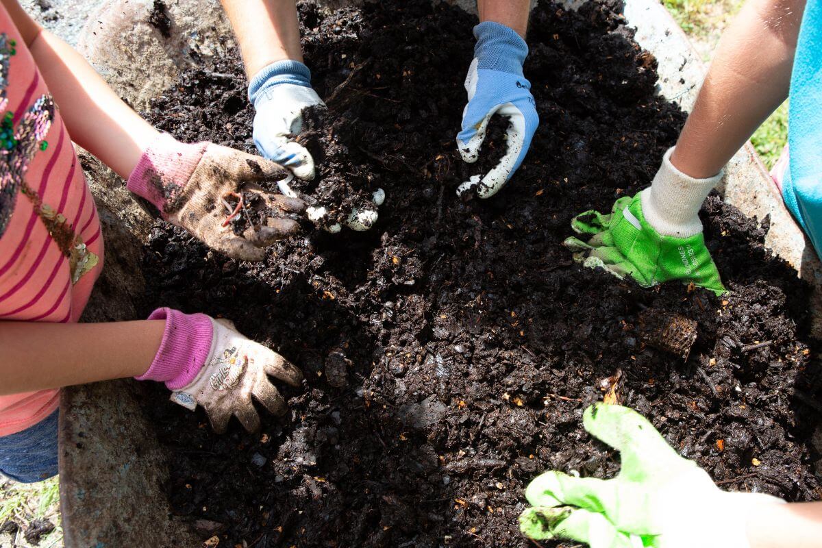 A group of kids with a parent, all wearing gloves, mixing soil and compost by hand in a garden.