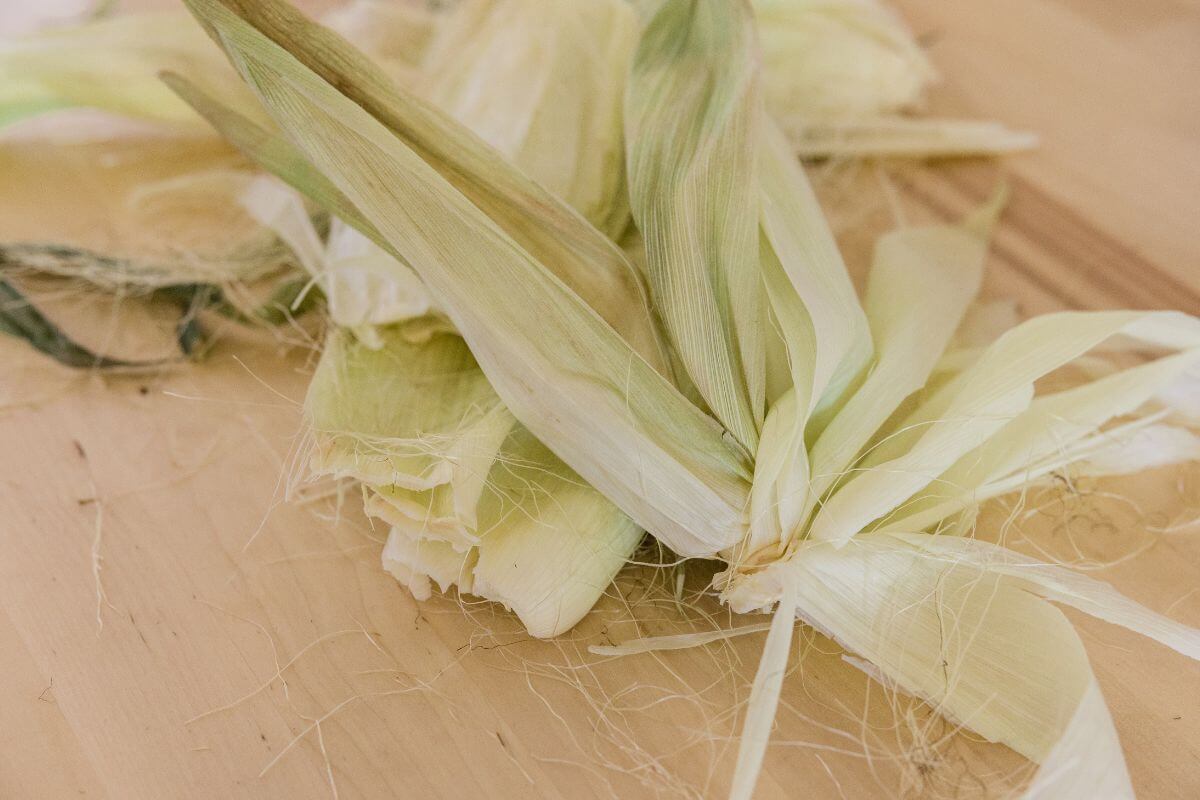 A fresh corn husks on a wooden surface. The leaves are light green and beige, with some husk threads visible. 