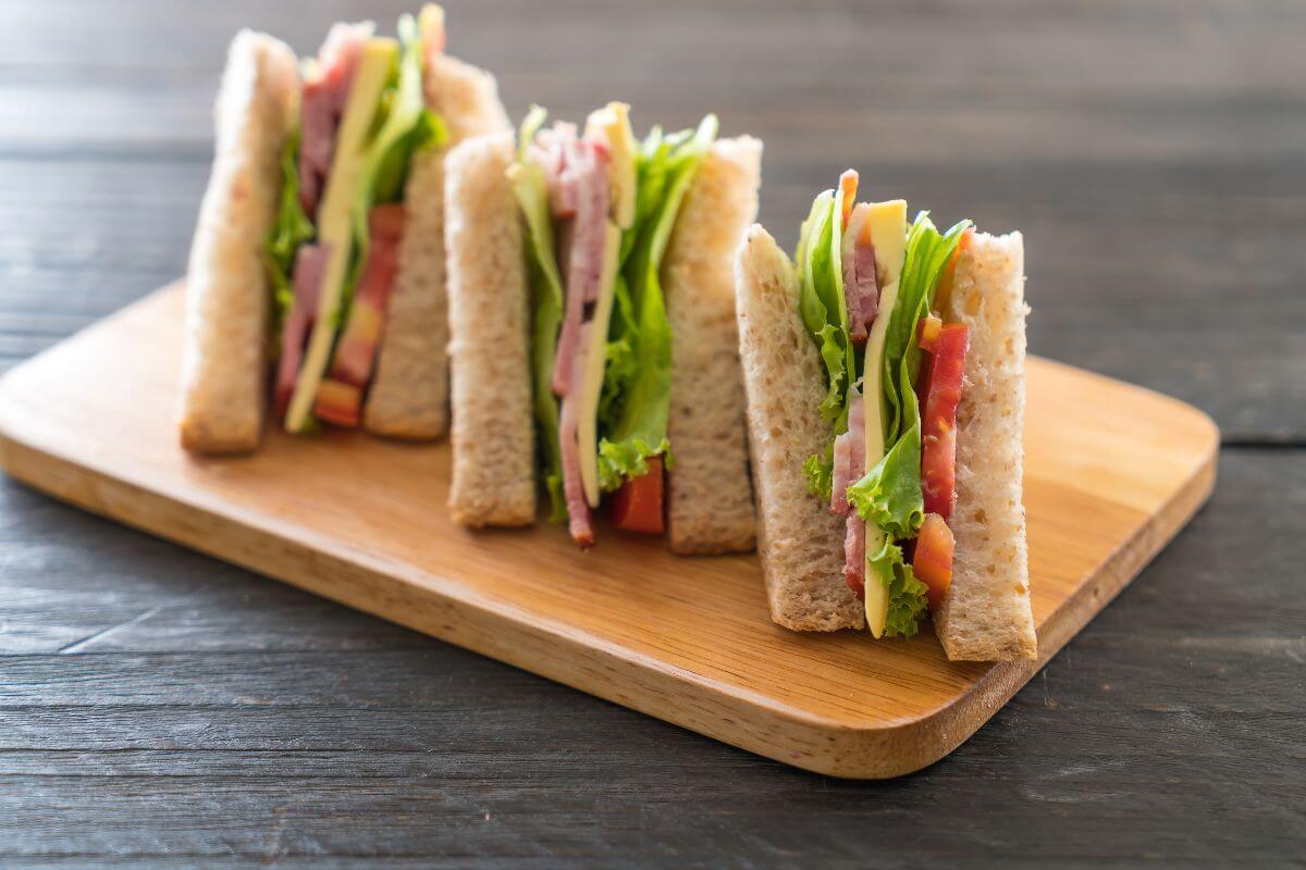 Four sandwich wedges filled with ham, lettuce, tomato, and cheese are neatly arranged on a rectangular wooden serving board. 