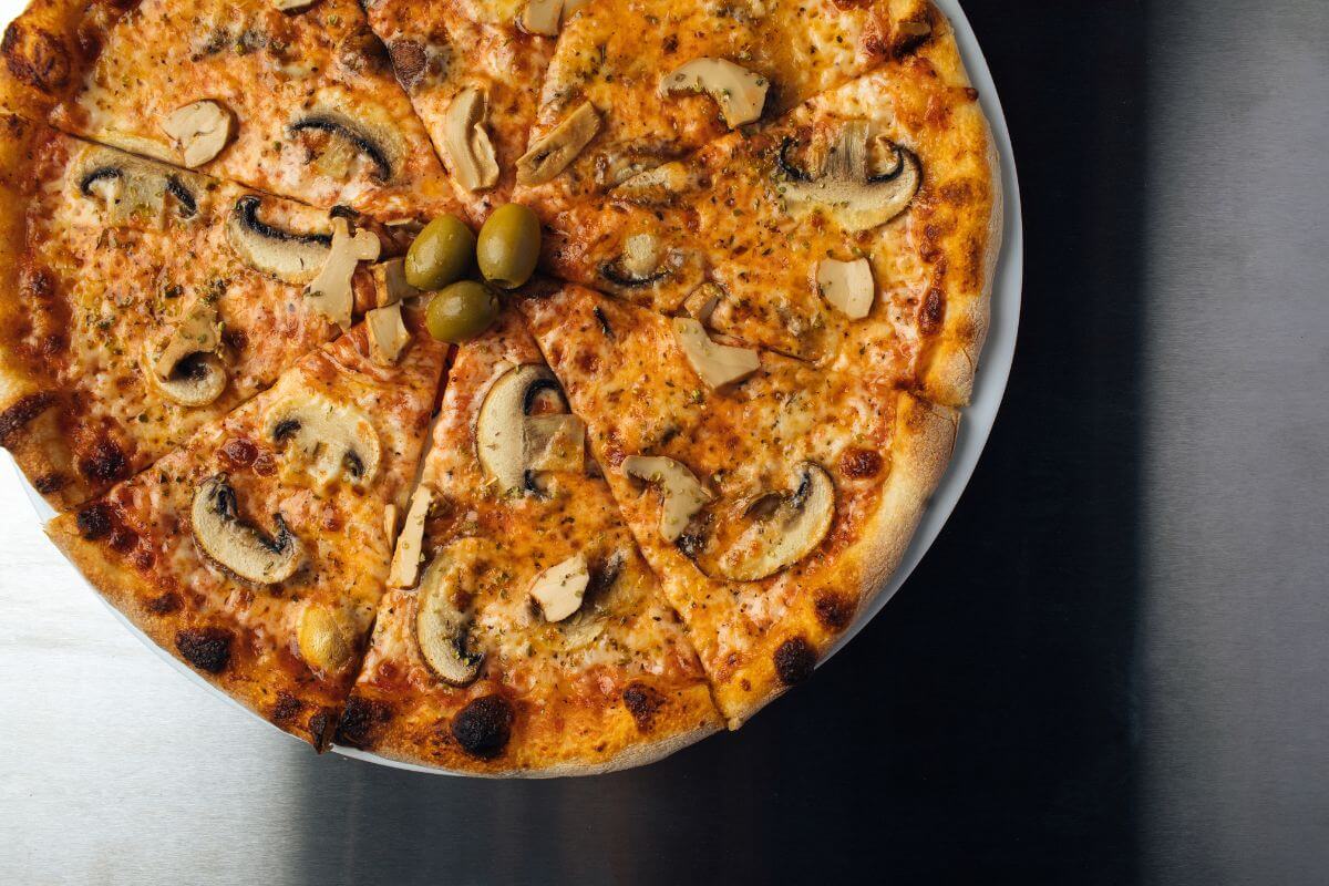 A whole pizza with a golden crust, topped with slices of mushrooms, chunks of artichoke hearts, and green olives. It is cut into eight slices and presented on a white plate against a metallic surface.