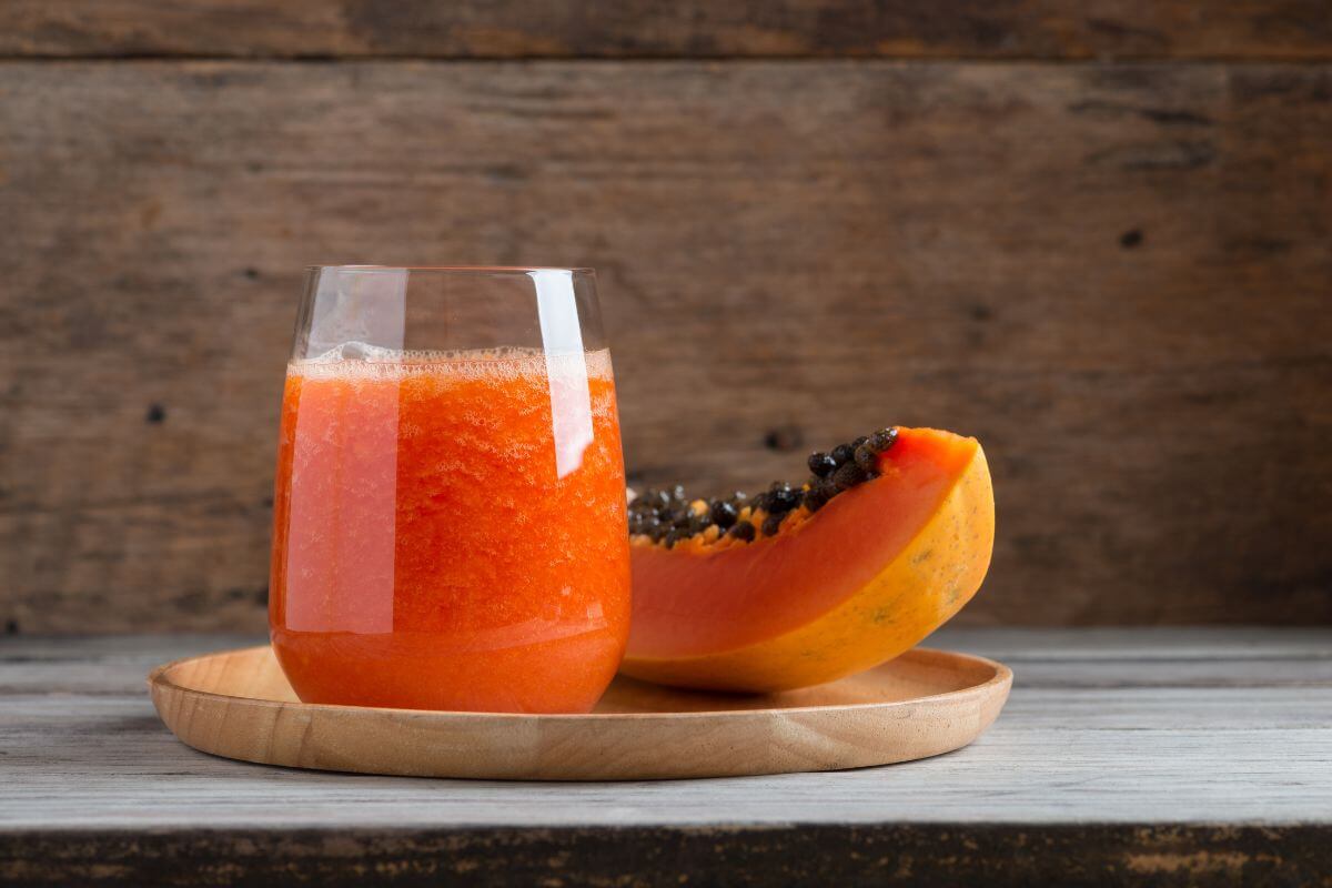 A glass filled with vibrant orange papaya smoothie is placed on a light wooden tray. Behind it, there is a half-cut papaya with black seeds, set against a rustic wooden backdrop. 