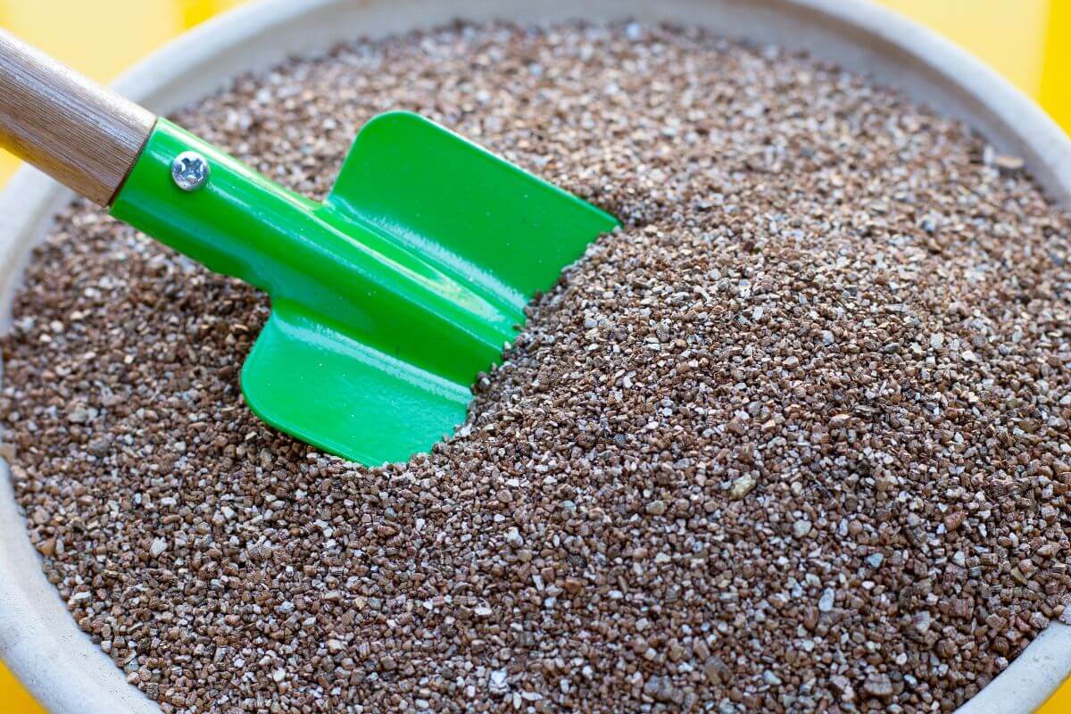 A close-up of a green-handled garden shovel resting in a pot filled with brown, finely granulated hydroponic vermiculite.