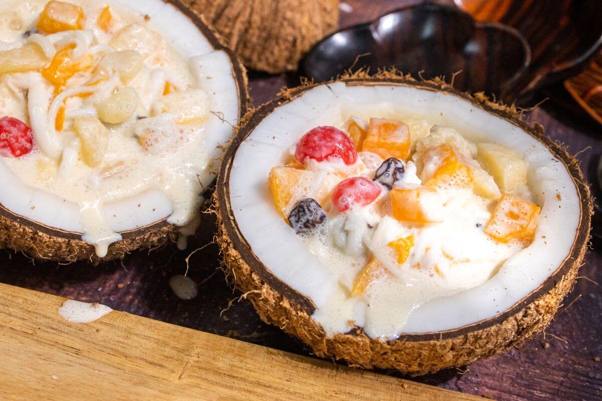 A creamy fruit salad served in halved coconut shells. The salad contains diced fruits such as papaya, grapes, and cherries, immersed in a creamy white dressing. 