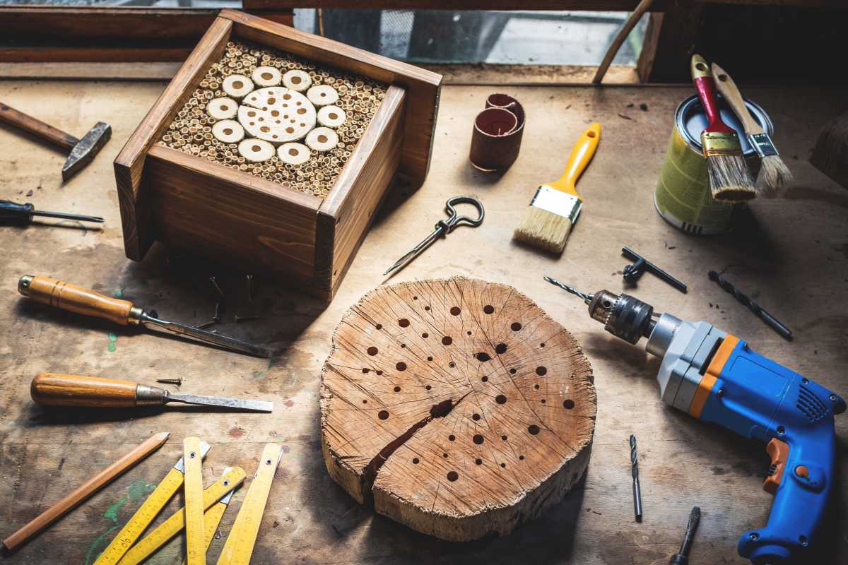 A workbench with various woodworking tools and materials, including a wooden block with drilled holes for a beneficial insect house.