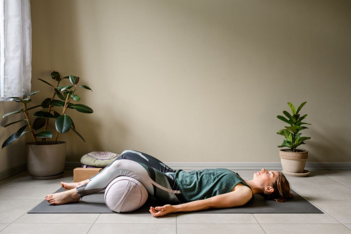 A person lying on a yoga mat in a well-lit room, relaxing in a reclined position with two potted plants are nearby.