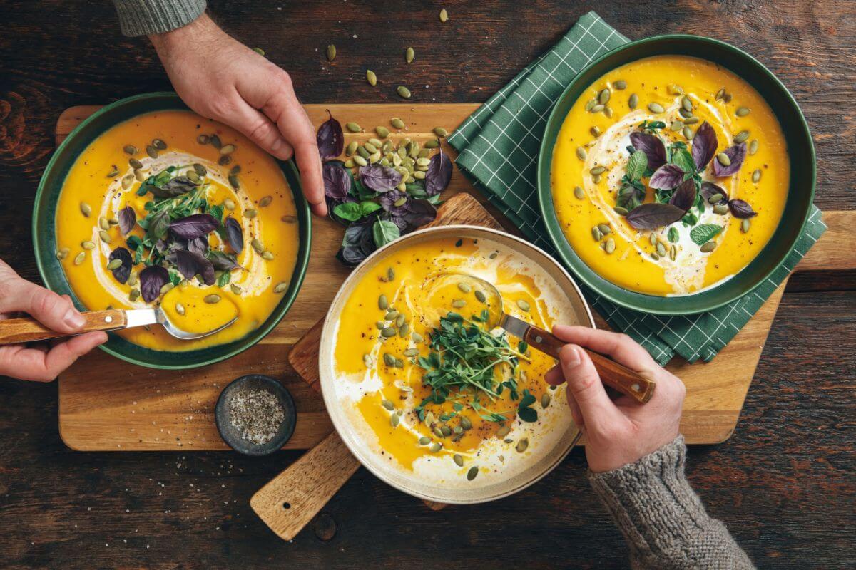 Three bowls of pumpkin soup are garnished with herbs and seeds, set on a wooden cutting board. 