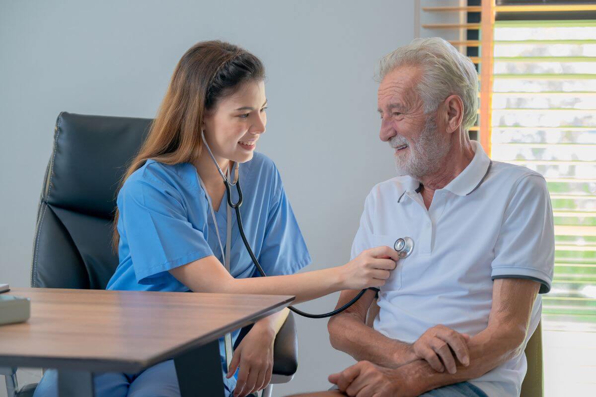 A female healthcare professional in blue scrubs uses a stethoscope to check the heart of an elderly male patient in a white polo shirt. 