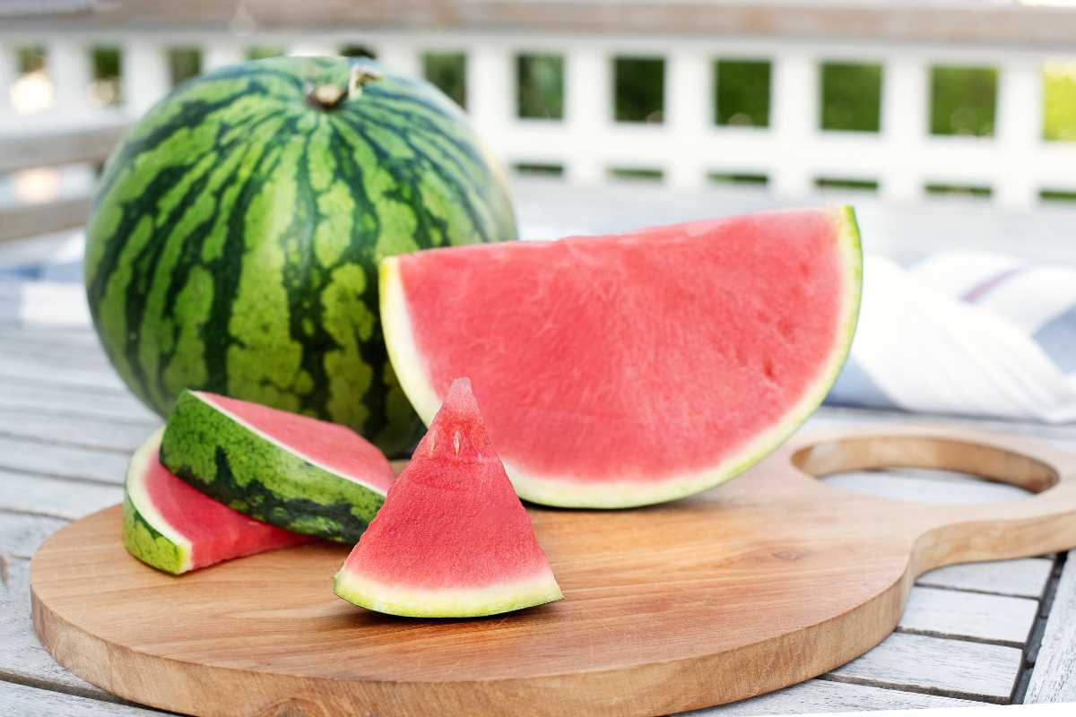 A round watermelon sits on a white wooden table with one large slice and two smaller slices placed on a wooden cutting board in front of it. 