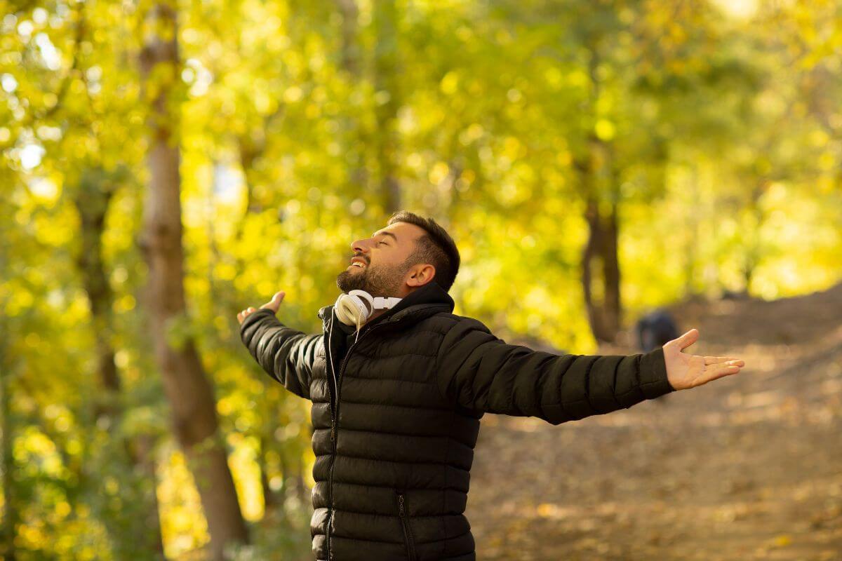 A person with a beard and short hair, wearing a black puffer jacket and headphones around their neck, is standing on a forest path engulfed in green and yellow foliage. 