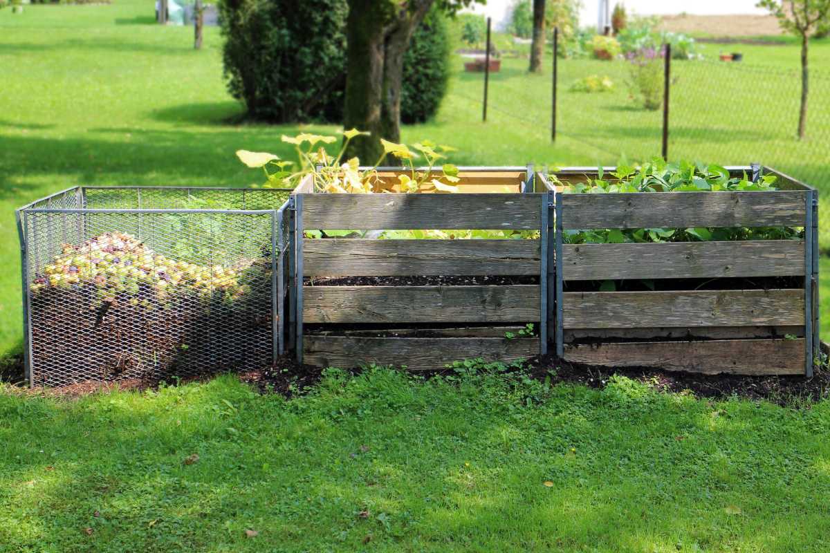 Three adjacent compost bins sit on a grassy lawn. The leftmost bin is made of wire mesh and is filled with decomposing organic matter.