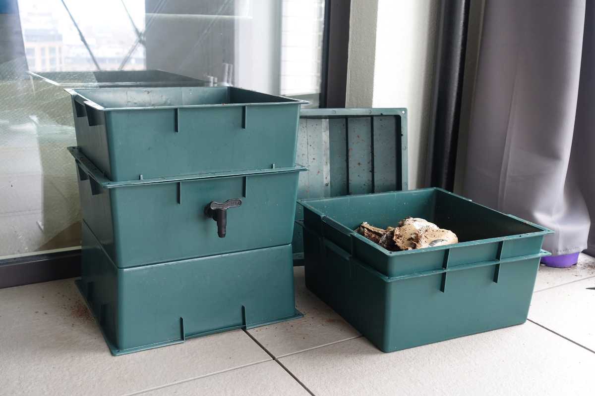 A stack of green, rectangular plastic worm bin sits on a tiled floor next to a window. One container on the right is open, revealing compost starter material inside.