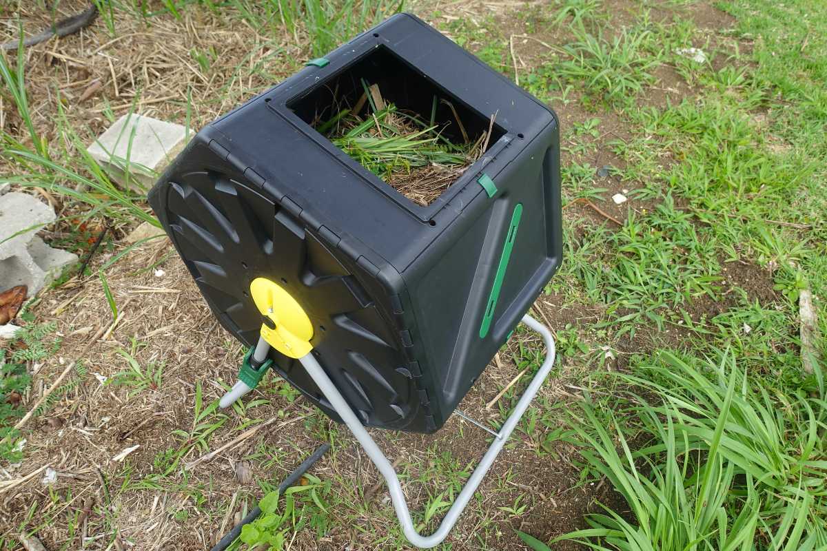 A black rotating tumbler compost bin, mounted on a metal stand, is situated outdoors on a patch of grass and soil.