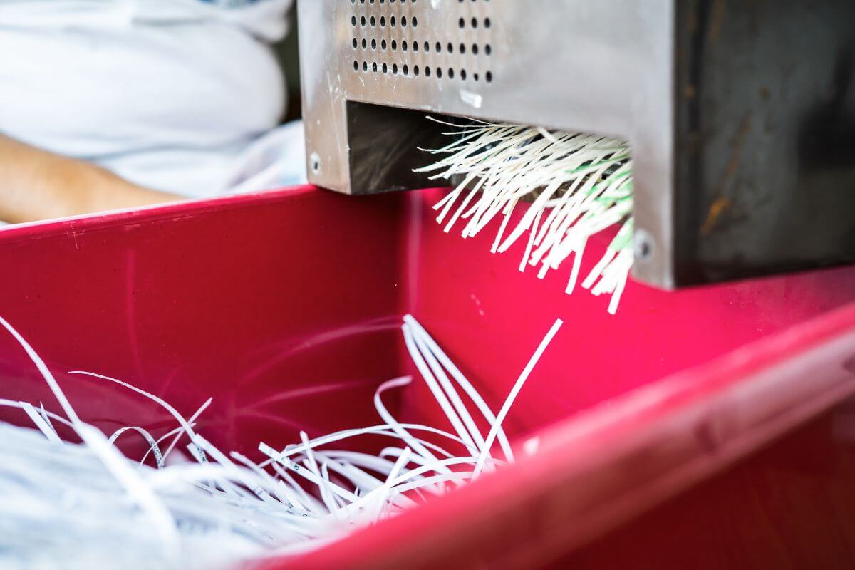 Close-up of a paper shredder shredding white paper into thin strips, which are falling into a red plastic bin below the shredder.