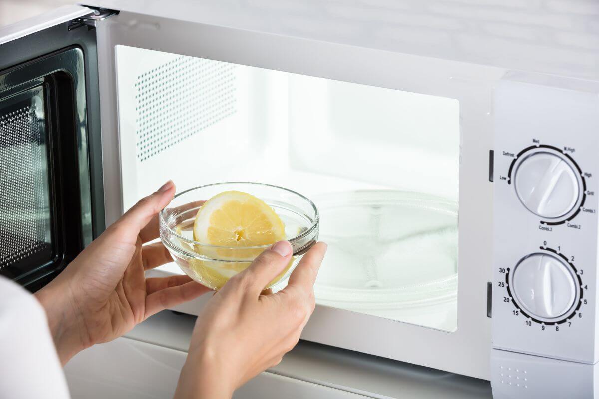A person holding a glass bowl with a halved lemon in front of an open microwave. The microwave, with two control dials on the right side, stands ready for use.