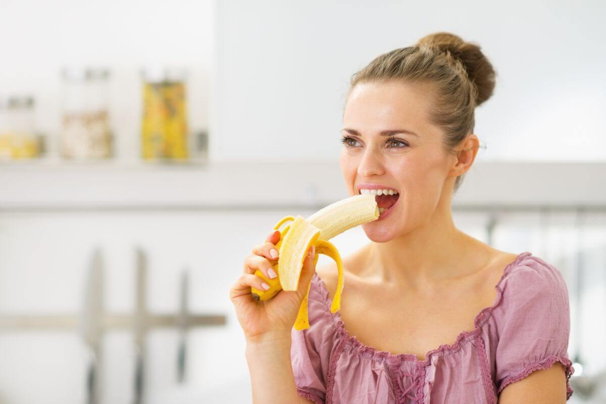 A woman with her hair tied up is standing in a kitchen and eating a peeled banana. 