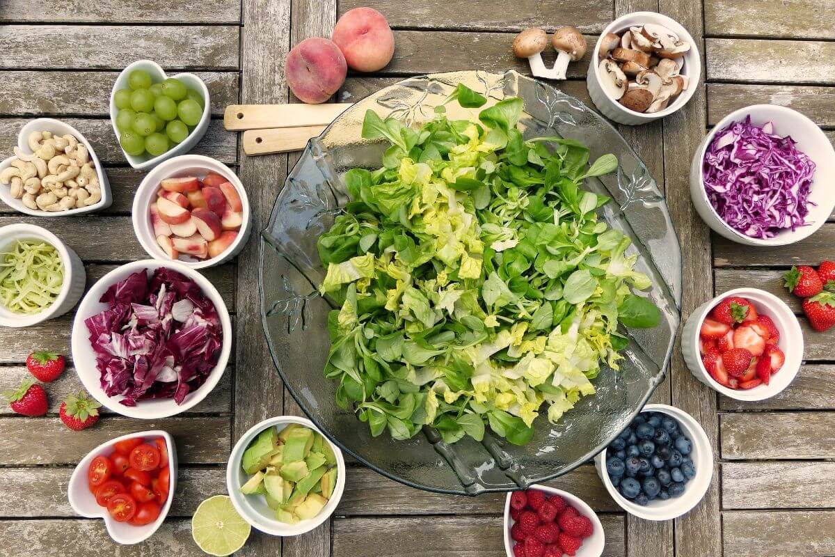 Centered on a wooden table is a large glass bowl filled with mixed green lettuce. Surrounding it are various smaller bowls containing an array of colorful ingredients, illustrating the contrast of fruit vs vegetable.