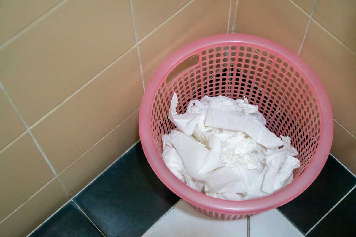 A pink plastic laundry basket filled with crumpled white paper towels, potential compost ingredients, sits in a corner of a tiled room with beige and dark green tiles on the wall and floor.