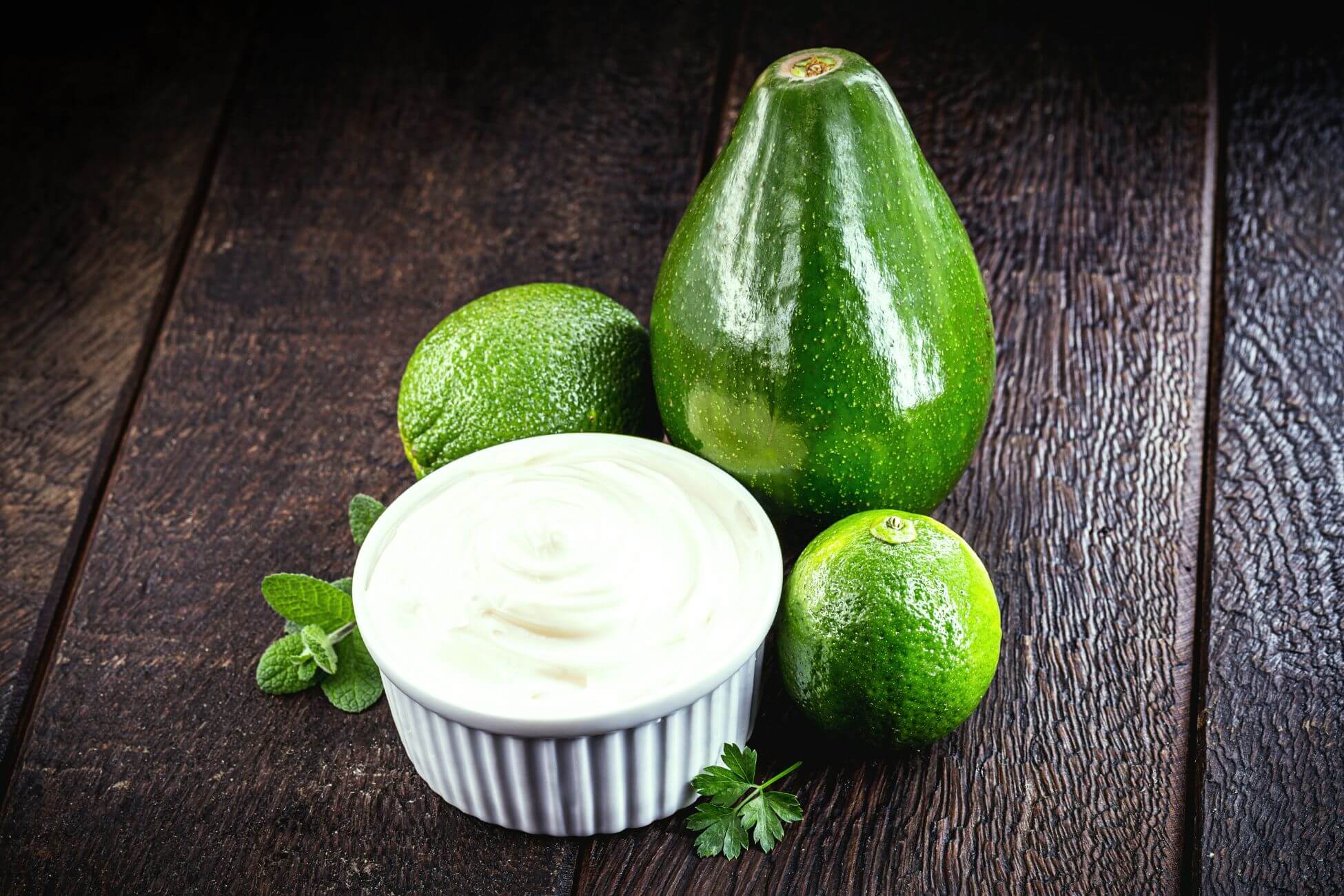 A bowl of creamy white avocado mayonnaise sits on a wooden surface alongside a whole avocado, two limes, and fresh mint and parsley leaves.