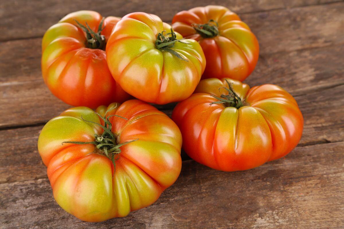 Five large, ripe hybrid tomatoes with red and green hues, ribbed texture, and glossy skin are arranged on a rustic wooden surface.