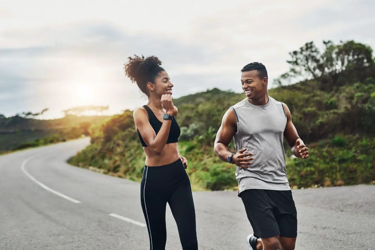 A man and woman jog together on a scenic road. She has a high ponytail and wears black athletic wear; he wears a gray tank top and black shorts. They smile and appear to be talking.