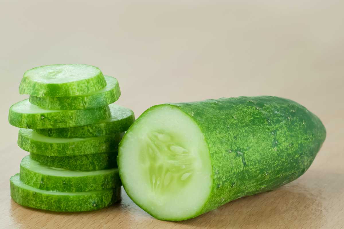 A sliced cucumber is displayed on a wooden surface. The cucumber is partially sliced, with several thin, round pieces stacked neatly beside the remaining uncut portion.