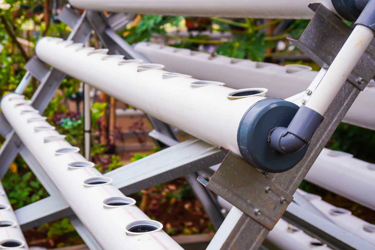 A vertical hydroponic farming system with white PVC pipes arranged diagonally. The pipes have round openings for planting and are supported by a metal frame.