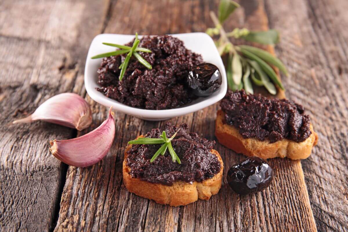A white dish filled with black olive tapenade garnished with rosemary sits on a rustic wooden surface. 