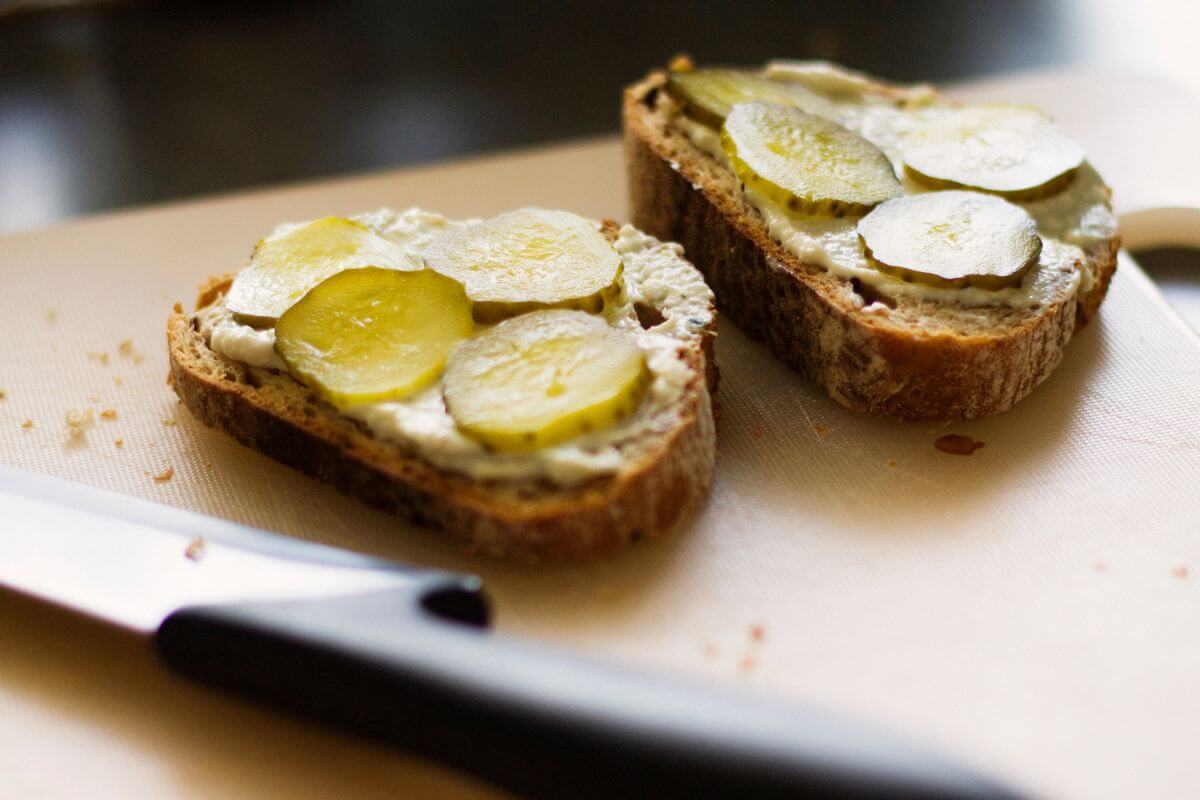 Two slices of bread with cream cheese and pickle slices placed on a cutting board.