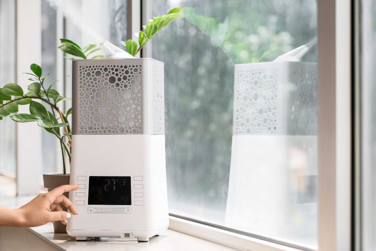 A white, modern humidifier sits on a windowsill next to green potted plants. A person's hand adjusts the settings on the digital control panel, which shows 27°C. 