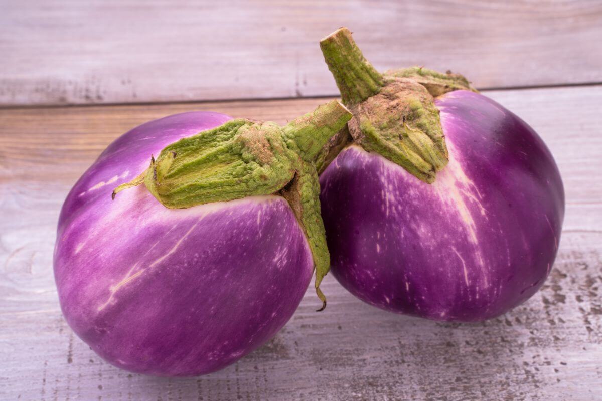 Two glossy Italian eggplants with green stems rest on a wooden surface, displaying light purple streaks on their skin.