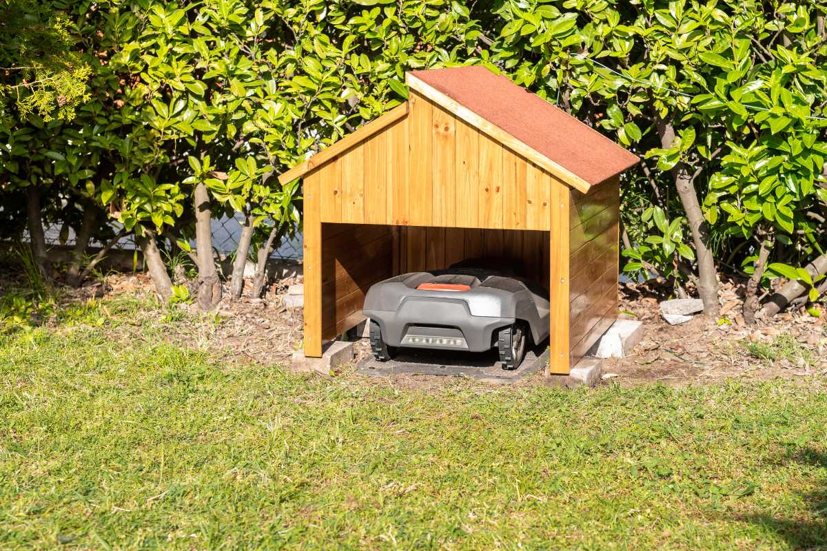 A robotic lawnmower is parked under a small wooden charging station with a red roof, situated in a garden near a hedge.