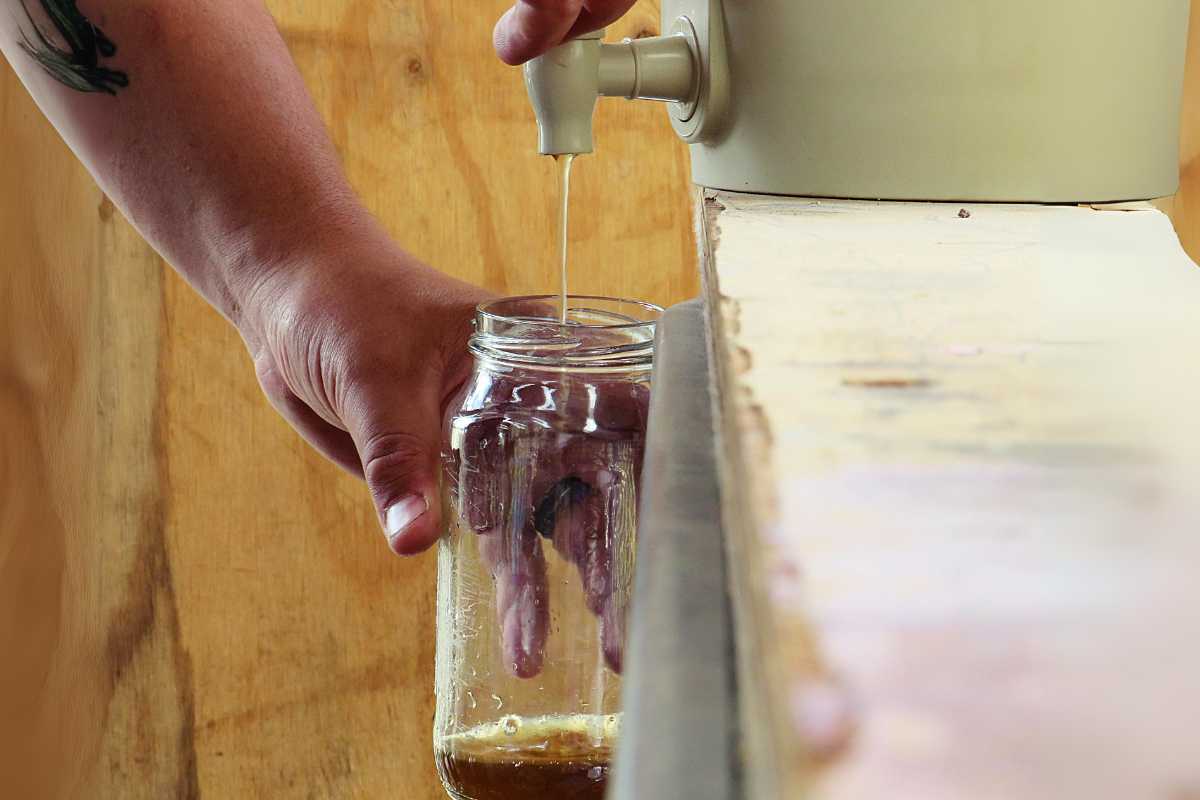 A person's hand is holding a mason jar under a tap, filling it with liquid. The background features a wooden surface on one side and a light-colored wall on the other. 