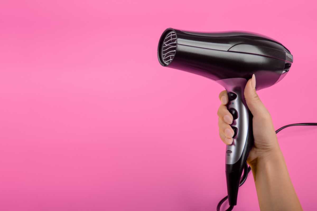 A hand holding a black hairdryer against a solid pink background, the hairdryer positioned with its nozzle facing left.