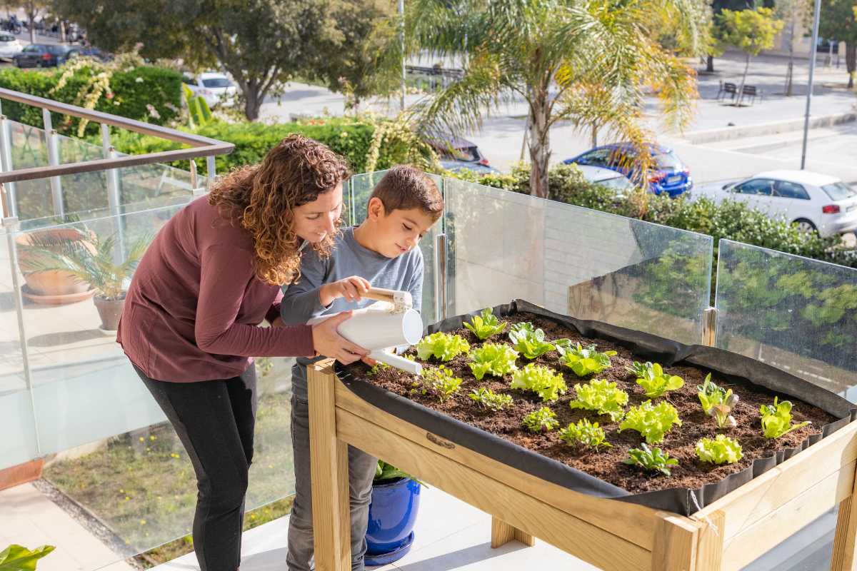 A woman and a child water plants in a raised wooden garden bed on a balcony, 