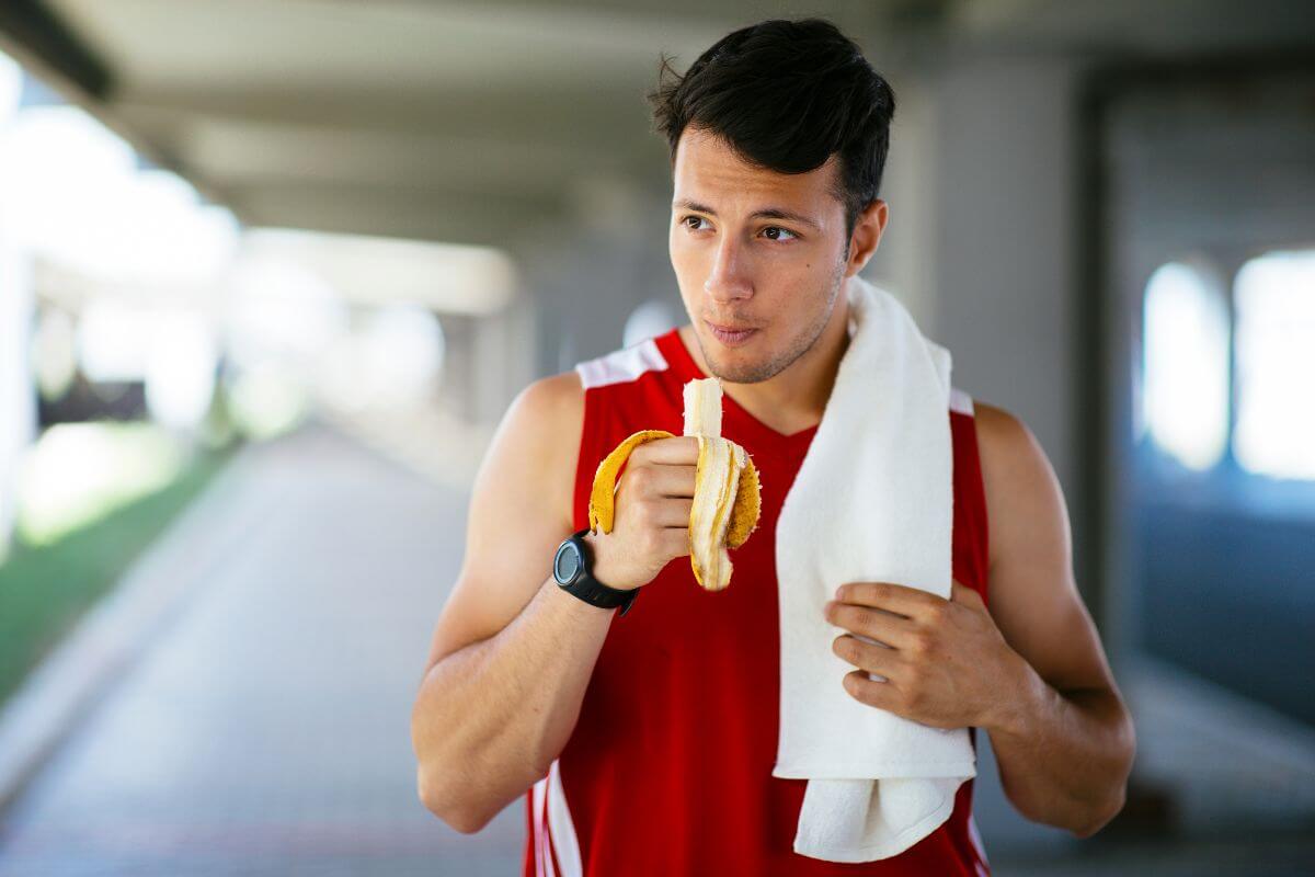 A man in a red sleeveless shirt, with a white towel draped over his shoulder, is eating a banana.