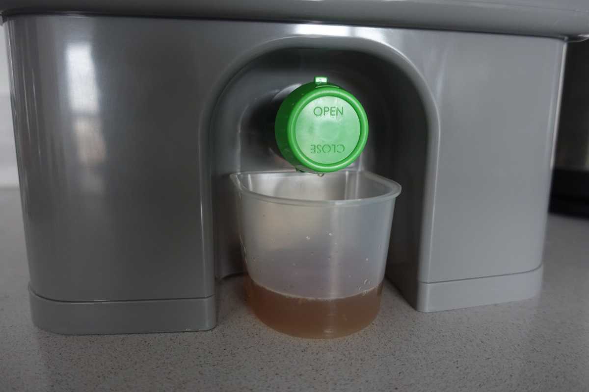 A bokashi compost bin with a green spout labeled "OPEN" and "CLOSE" dispenses a brown liquid into a clear plastic cup placed underneath. 