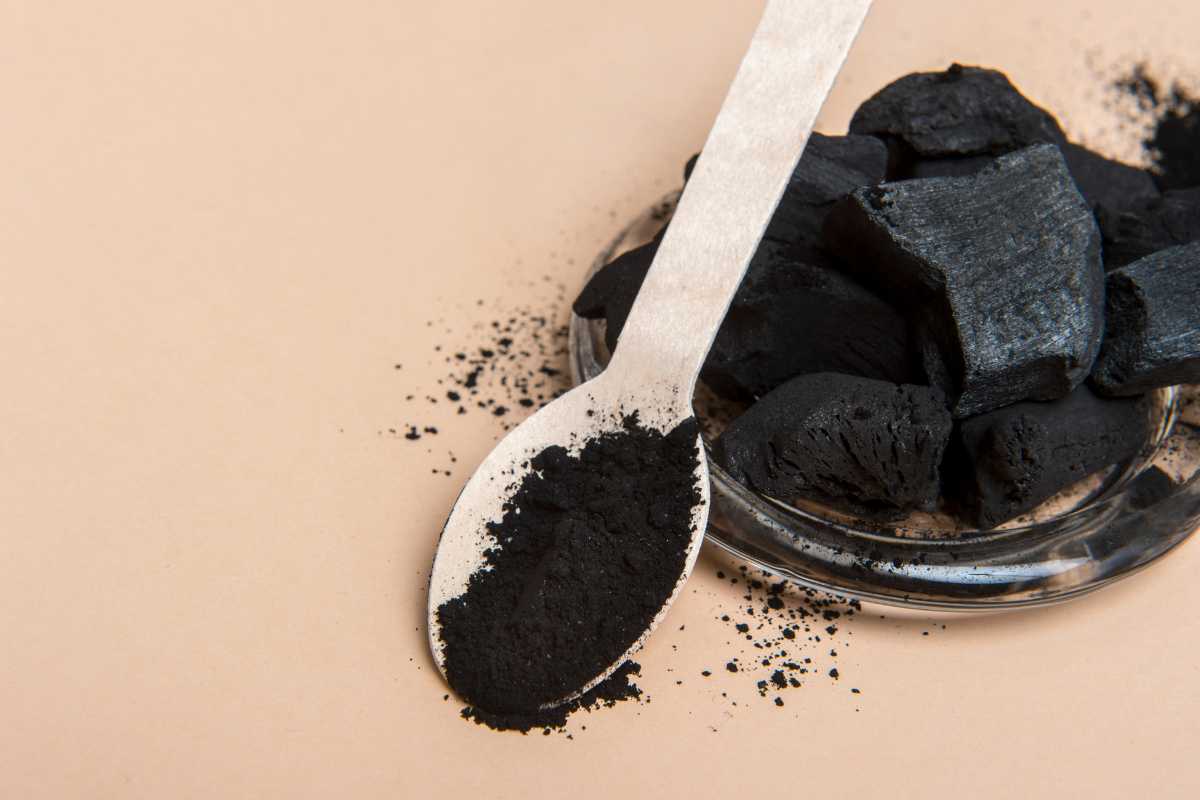 A metal spoon containing powdered activated charcoal next to a small glass plate filled with chunks of activated charcoal on a beige background. 