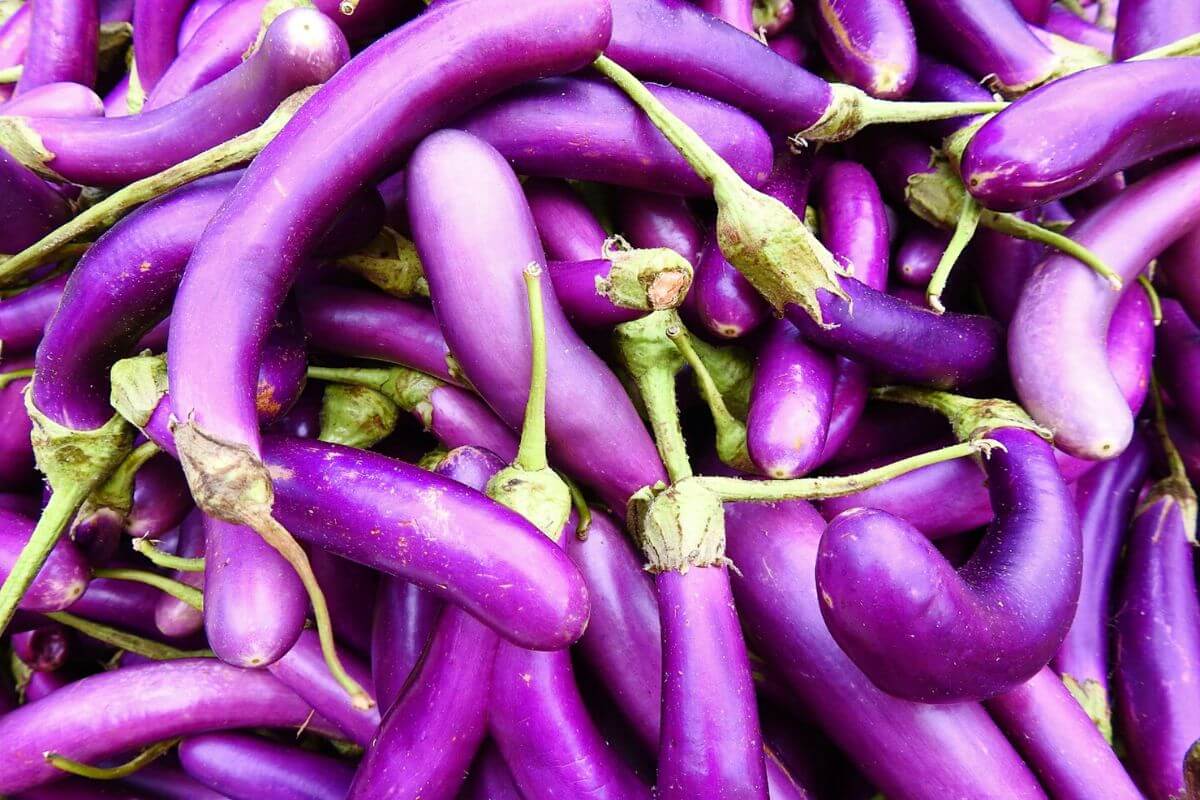 A close-up of a vibrant pile of long, slender purple Chinese eggplants, some with stems and leaves attached. 