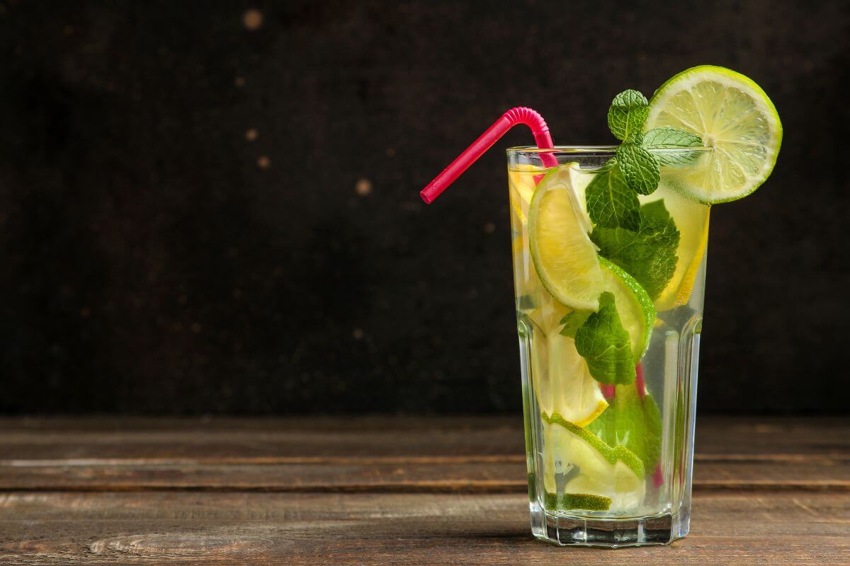 A tall glass of refreshing lemonade with ice, garnished with slices of lime and fresh mint leaves. A red straw is placed in the drink. 