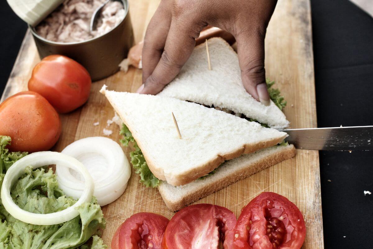 A hand holds two triangular sandwich halves made of white bread, with lettuce, onion, and possibly other fillings, while cutting the sandwich with a knife. 