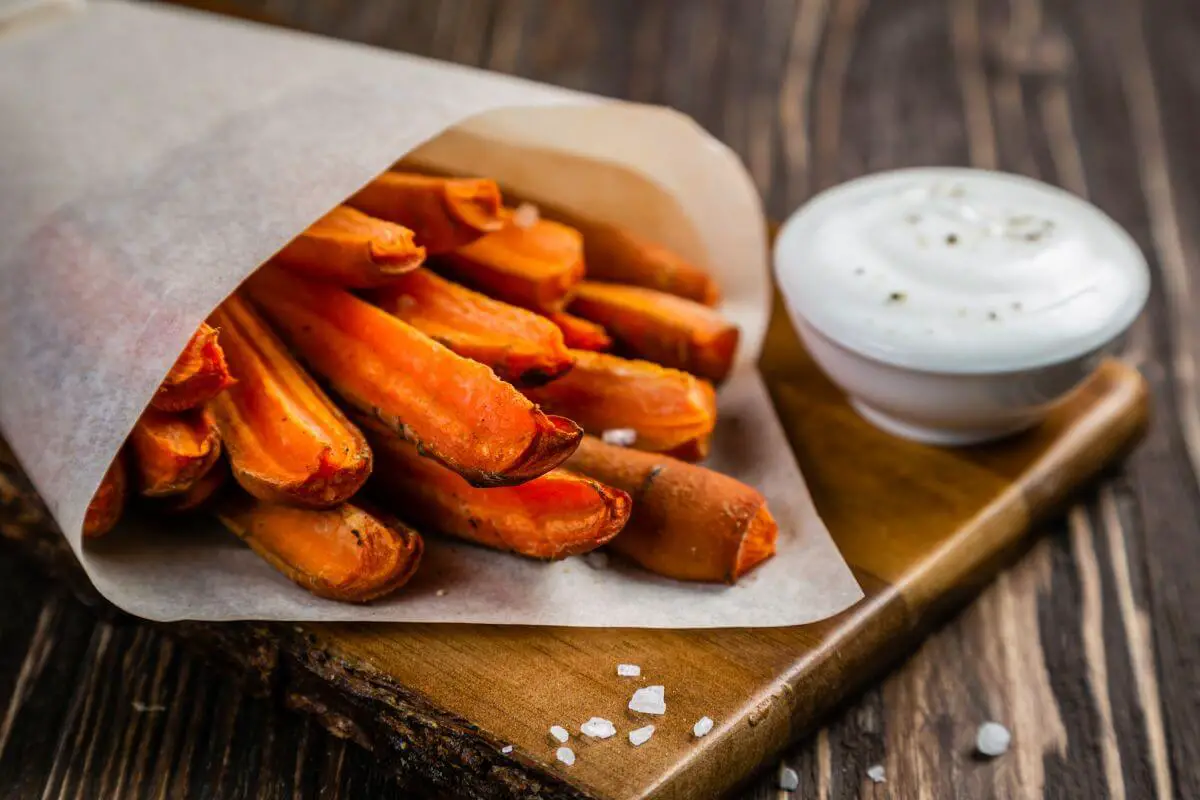 A bundle of baked carrot fries wrapped in parchment paper rests on a wooden board, with a bowl of creamy white dipping sauce garnished with black pepper nearby. Coarse salt is scattered on the rustic wooden surface.