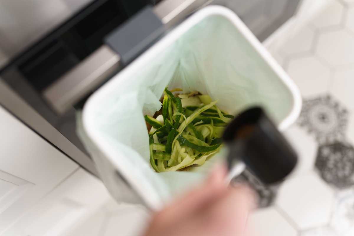 A hand is seen discarding sliced vegetable peels into a white bokashi compost bin lined with a green plastic bag.