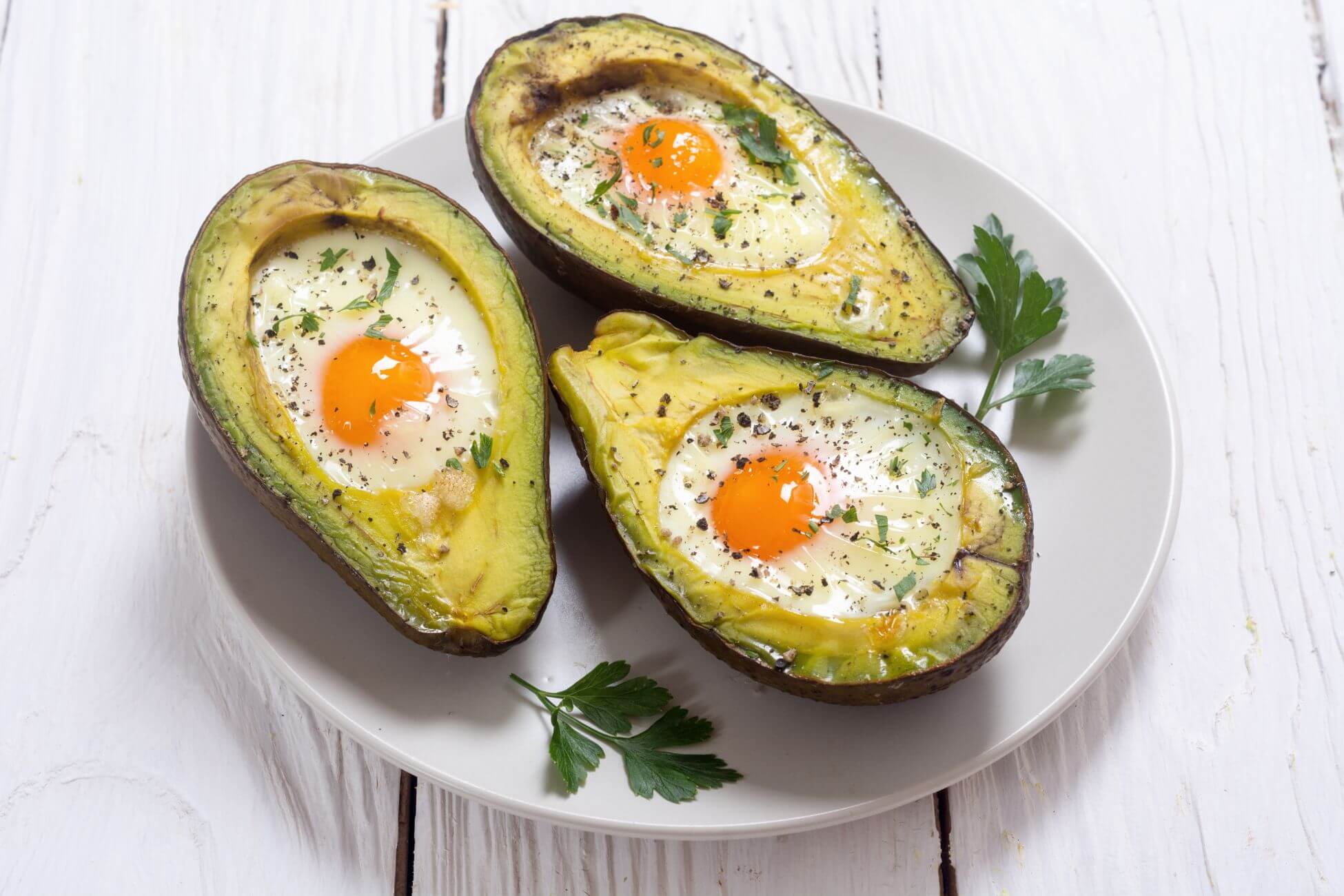 Half of an baked avocado with the pit removed, and an egg cooked sunny-side-up in each hollow sits on a white plate. 